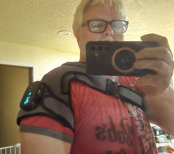 Older woman with glasses taking a selfie while wearing a black shoulder heating massager on her right shoulder