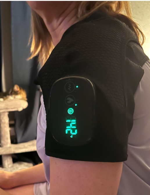 Close-up of woman wearing black shoulder massager with illuminated digital display showing temperature and control buttons